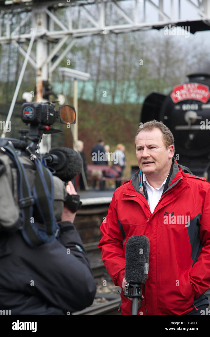 Sky tv reporter holding microphone and delivering piece to camera in front of blurred flying Scotsman, east lancashire railway in bury lancashire uk Stock Photo