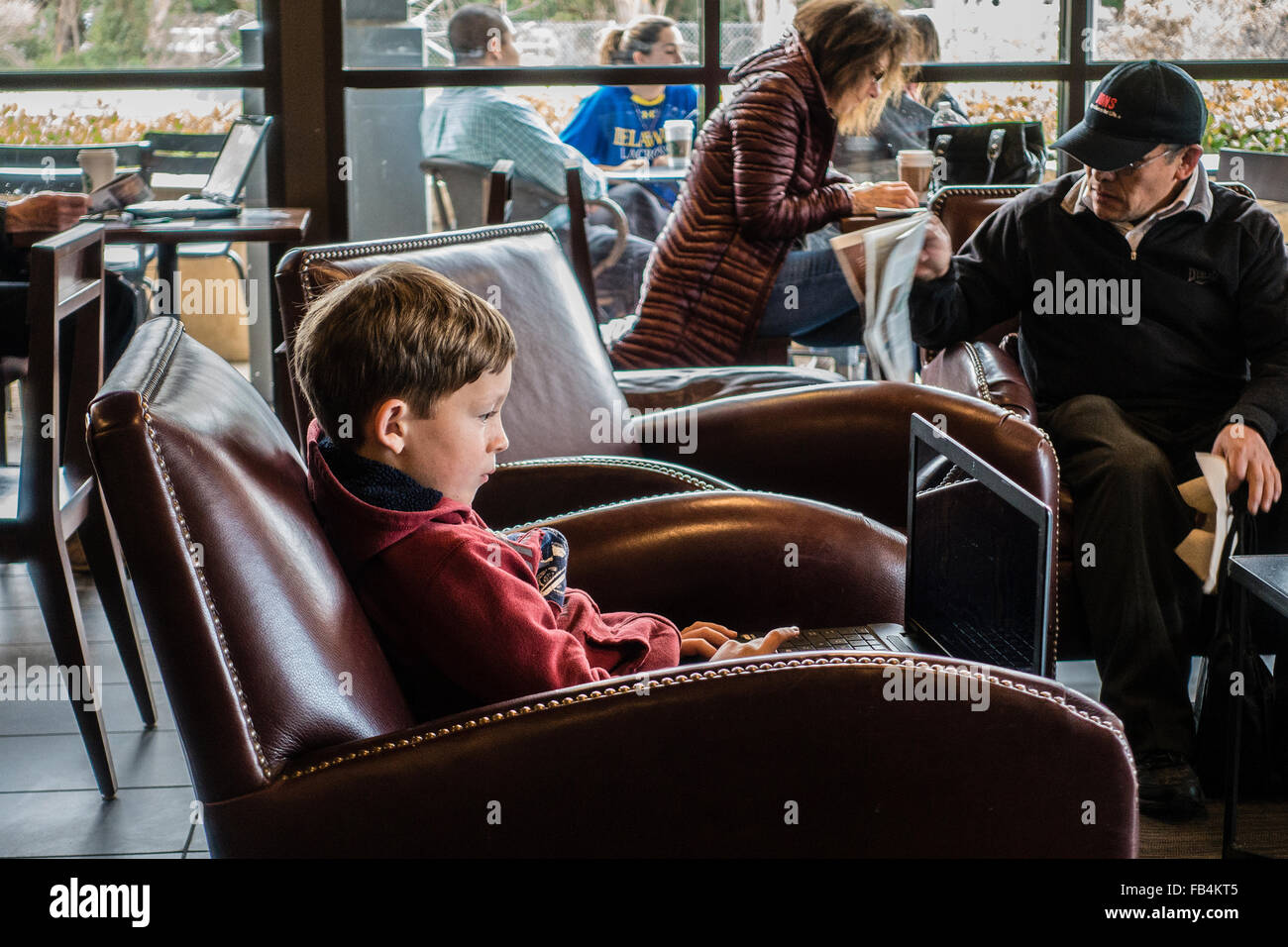 A young technically savvy boy, 10-12 years old, sits in a coffee shop with a big laptop on his lap as he concentrates on it. Stock Photo
