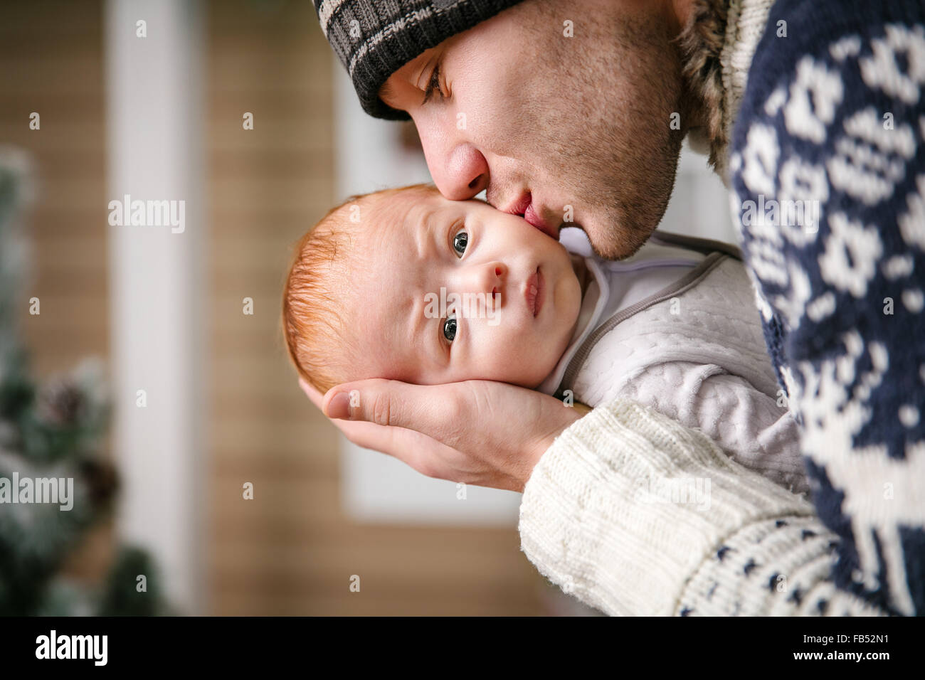 Father holding and kissing baby son among while wearing winter hat, with Christmas tree in background Stock Photo