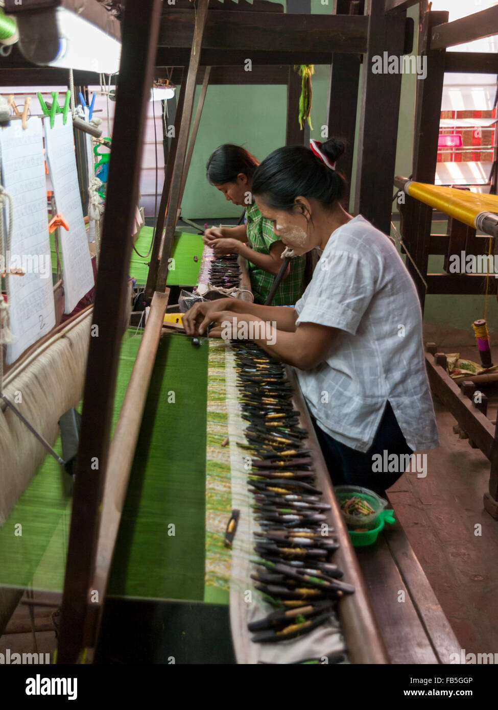 Two women weaving by using shuttles with thread spools on a loom. Partially woven green silk fabric visible. Mandalay, Myanmar. Stock Photo