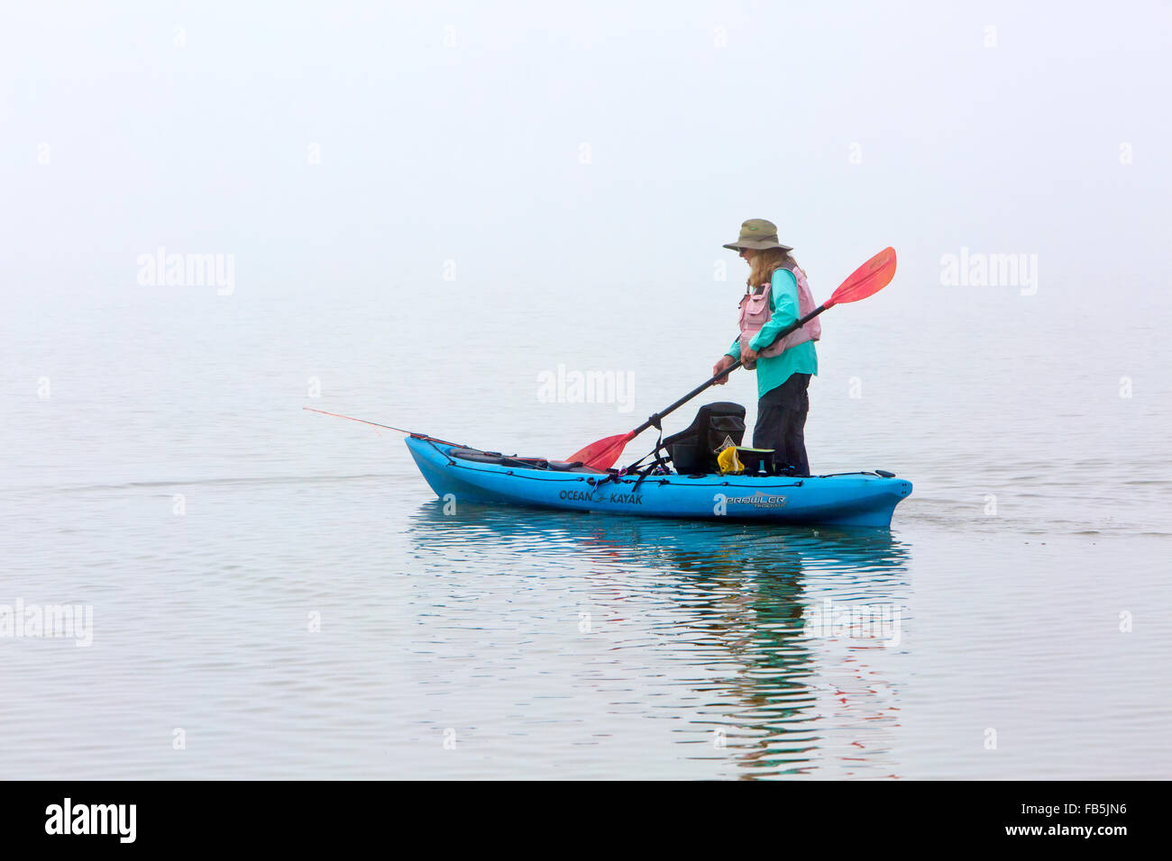 Woman maneuvering ocean kayak containing fishing gear, foggy coastal bay. Stock Photo