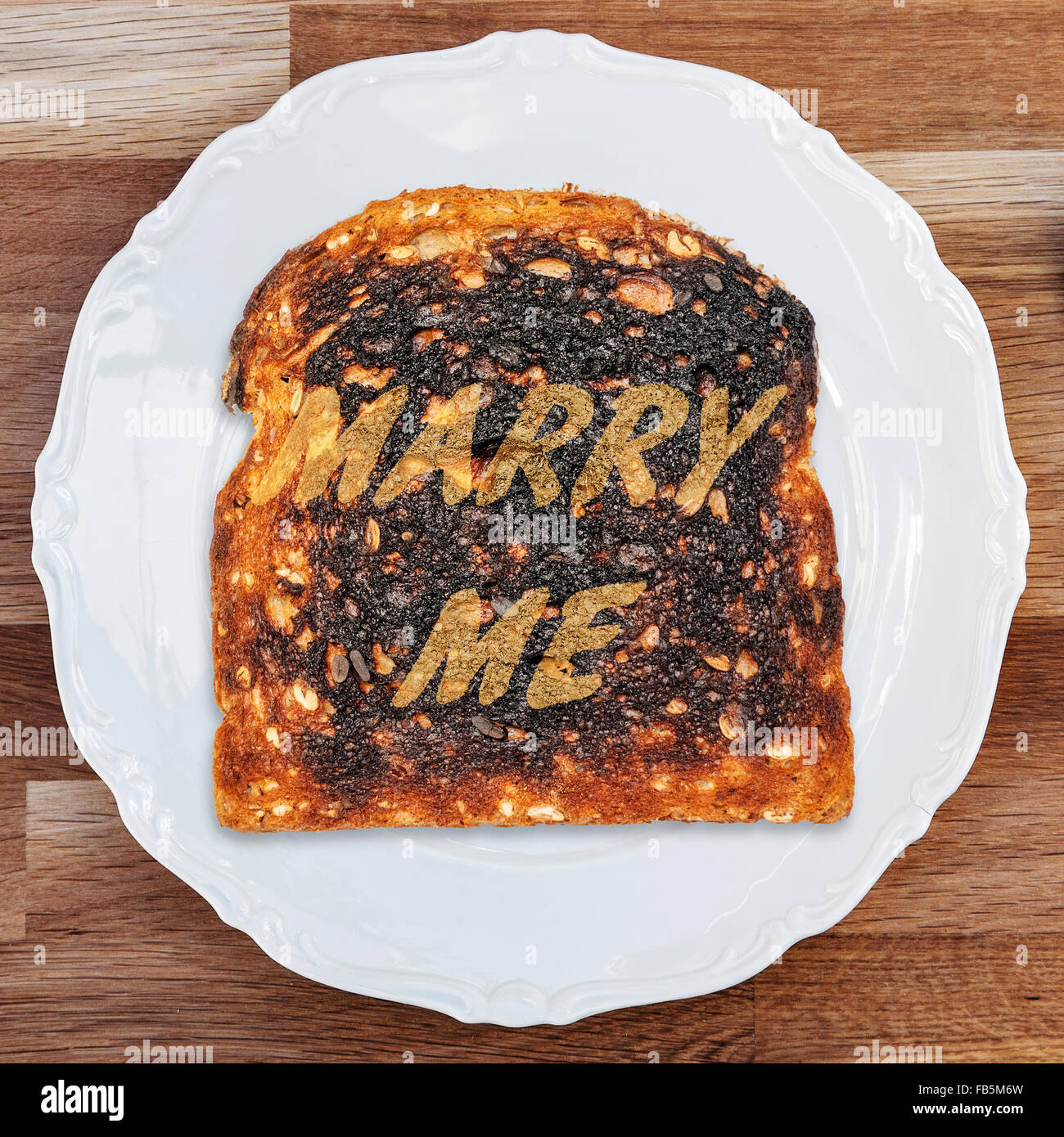A slice of burnt toast with a marraige proposal message on a shiny white plate placed upon a wooden table surface. Stock Photo
