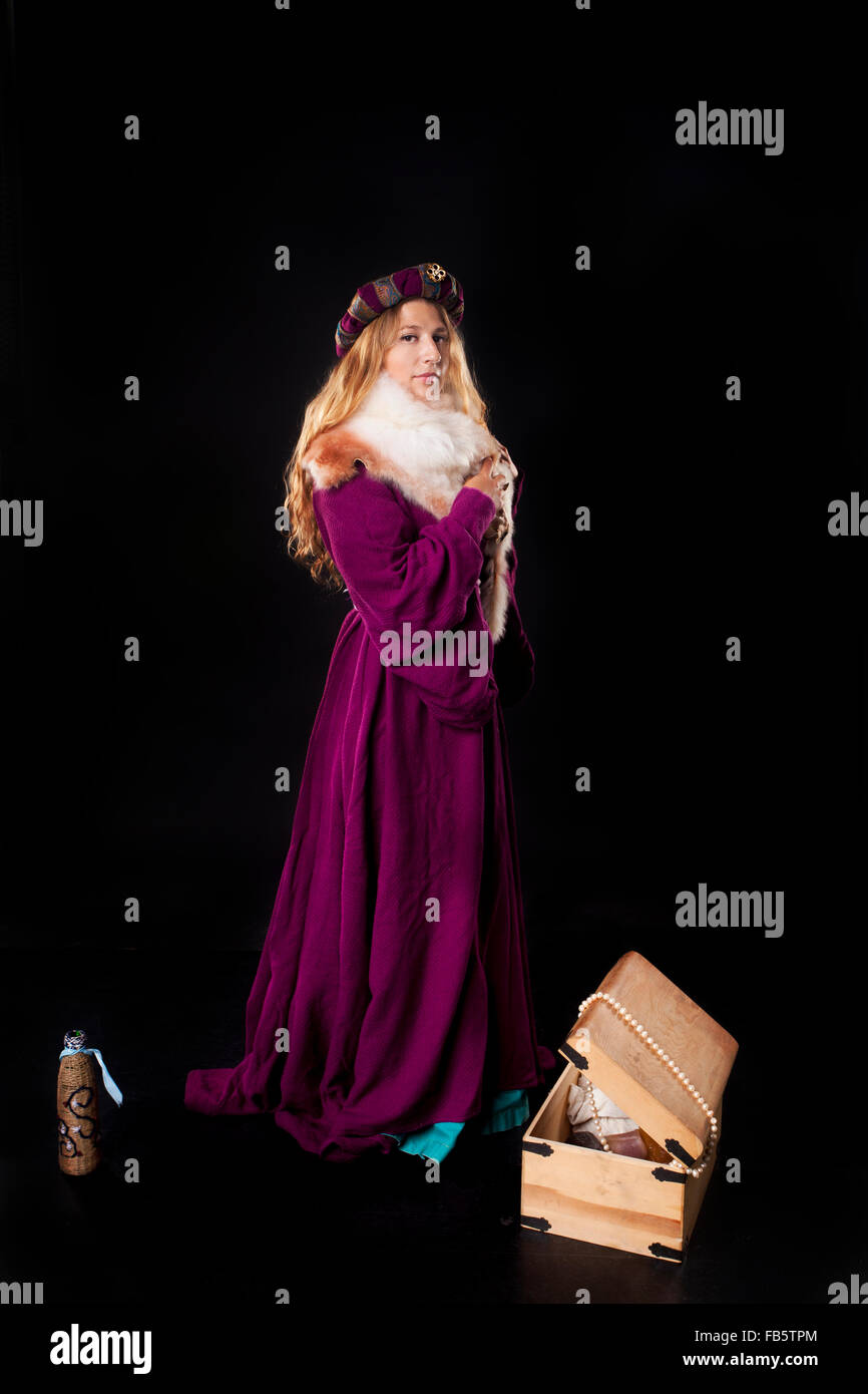 Studio portrait of beautiful girl dressed as a medieval noble lady in purple mantle and fur with a treasure chest on black backg Stock Photo