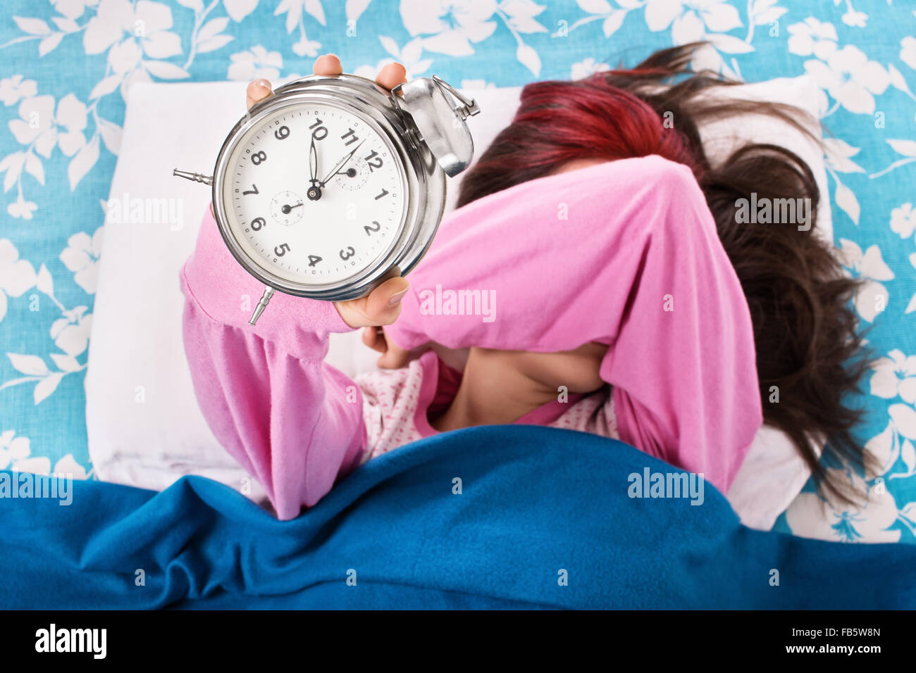 Young woman lying in bed covering her eyes with her arm, holding an alarm clock. Student doesn’t want to wake up early for university or school. Stock Photo