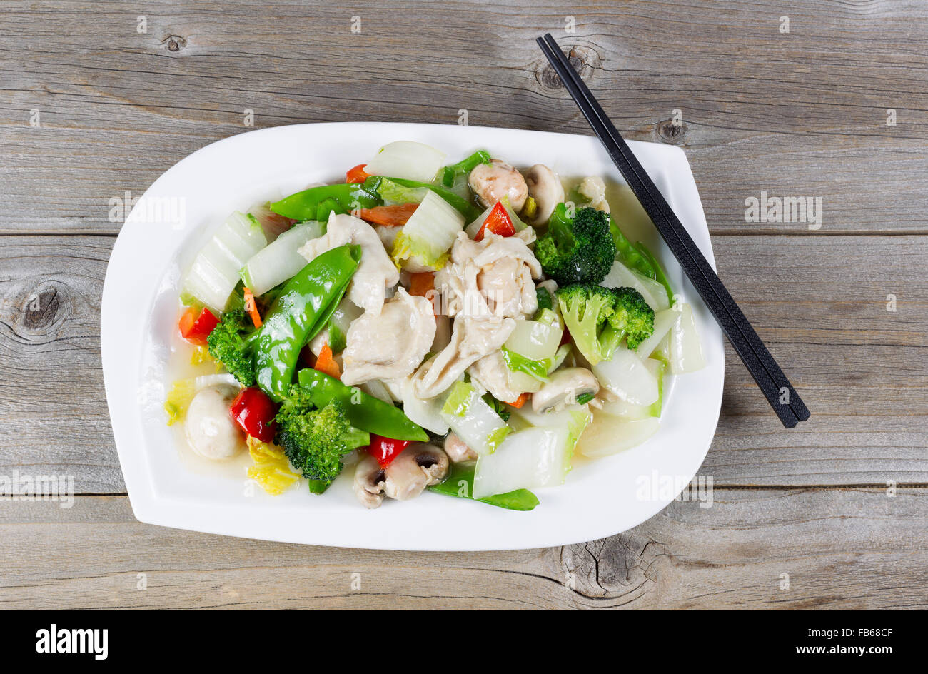 Top view of stir fried white chicken pieces with broccoli, snow peas, peppers and mushroom in white plate on rustic wood setting Stock Photo