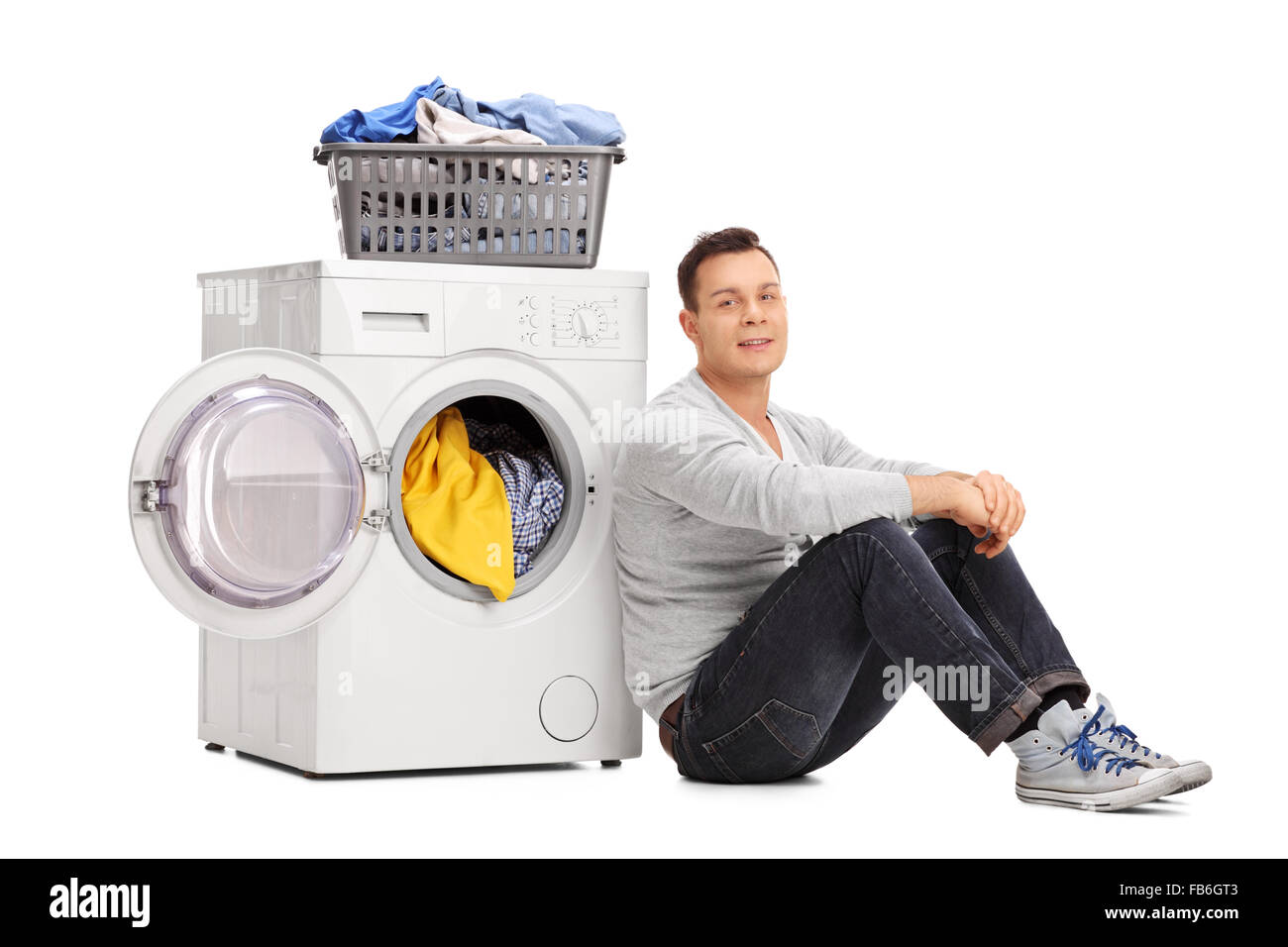Young man sitting by a washing machine and looking at the camera isolated on white background Stock Photo
