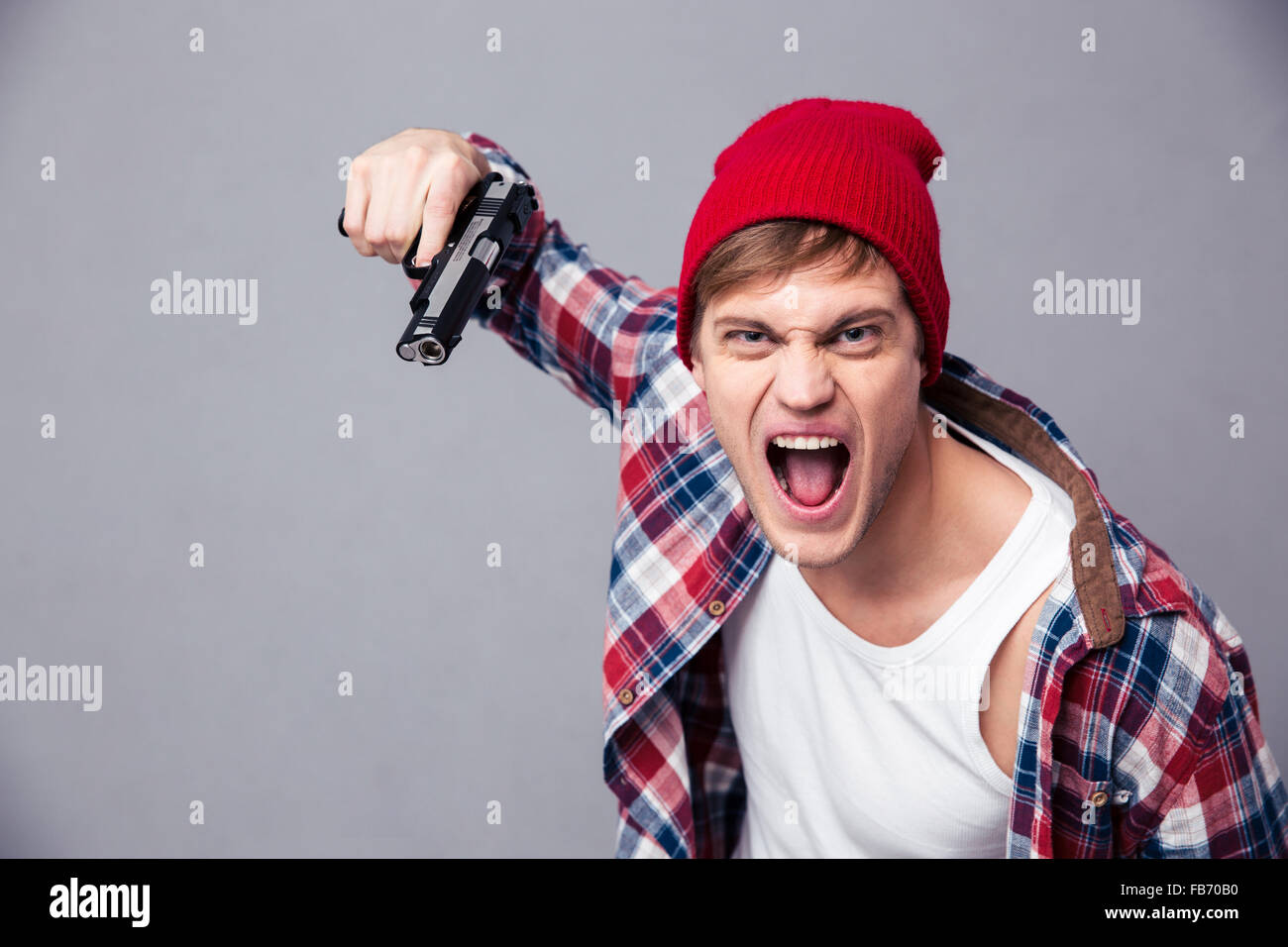 Dangerous agressive young man in checkered shirt and red hat shouting and threatening with gun over grey background Stock Photo