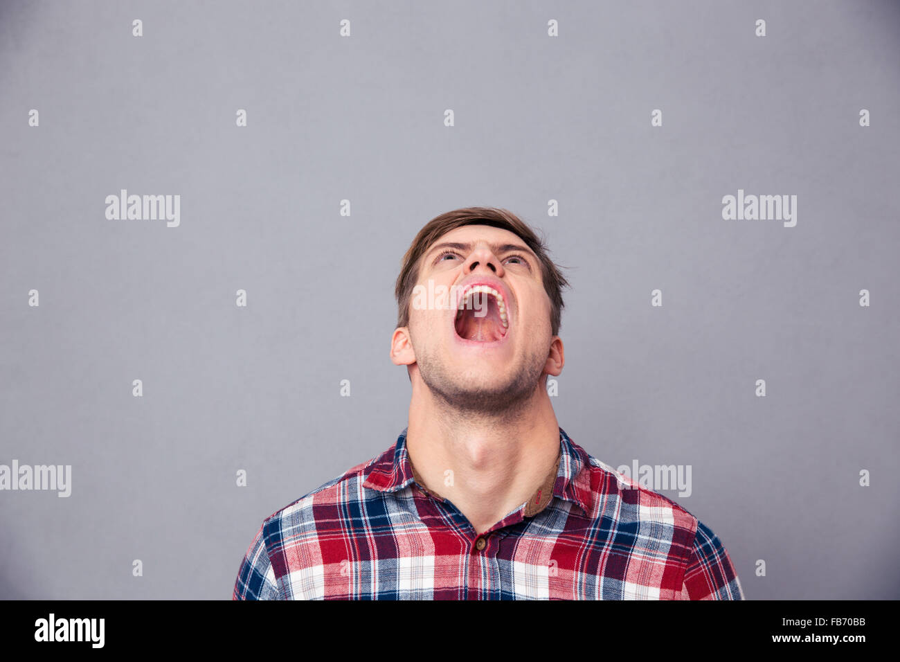 Despaired angry young man in plaid shirt looking up and screaming over grey background Stock Photo