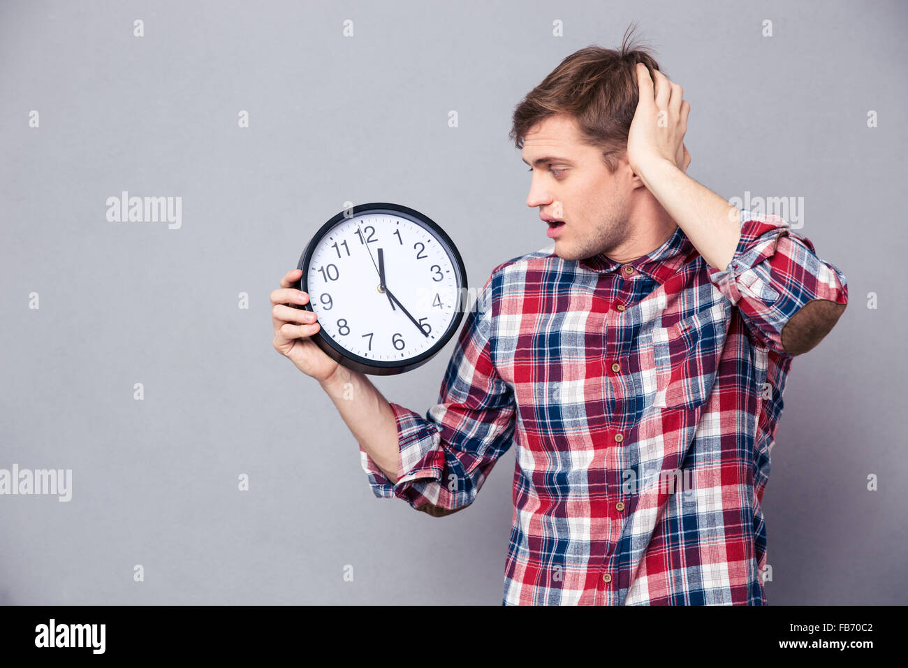 Worried amazed young man in checkered shirt holding and looking at clock over grey background Stock Photo