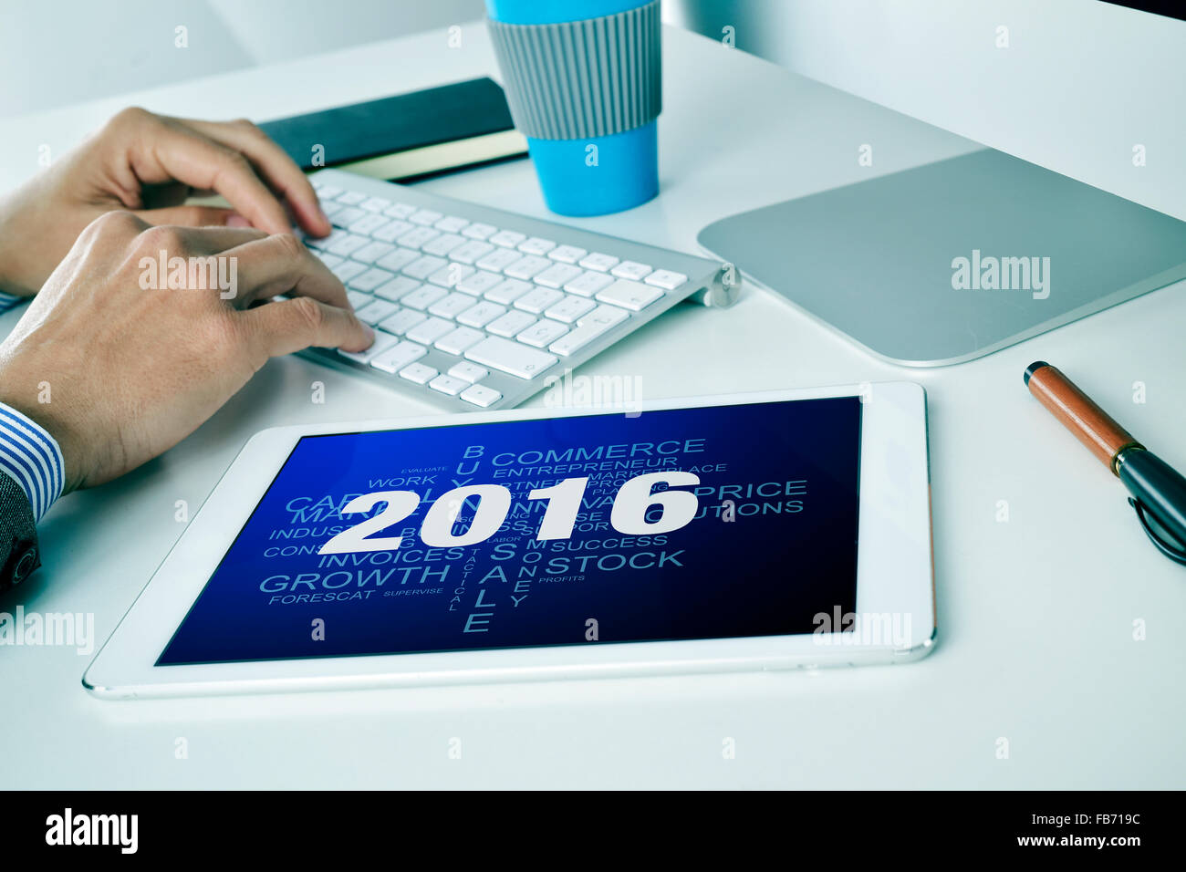 a young caucasian businessman sitting at his office desk where there is a tablet computer with a tag cloud of goals for the 2016 Stock Photo