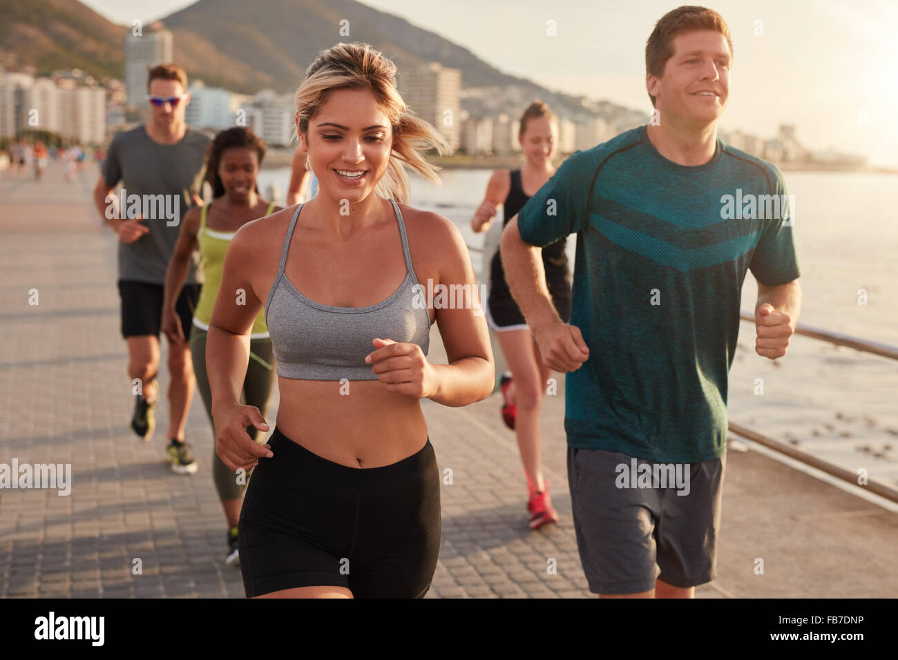 Portrait of fit young woman running with friends on street along the sea. Running club group training outdoors. Stock Photo