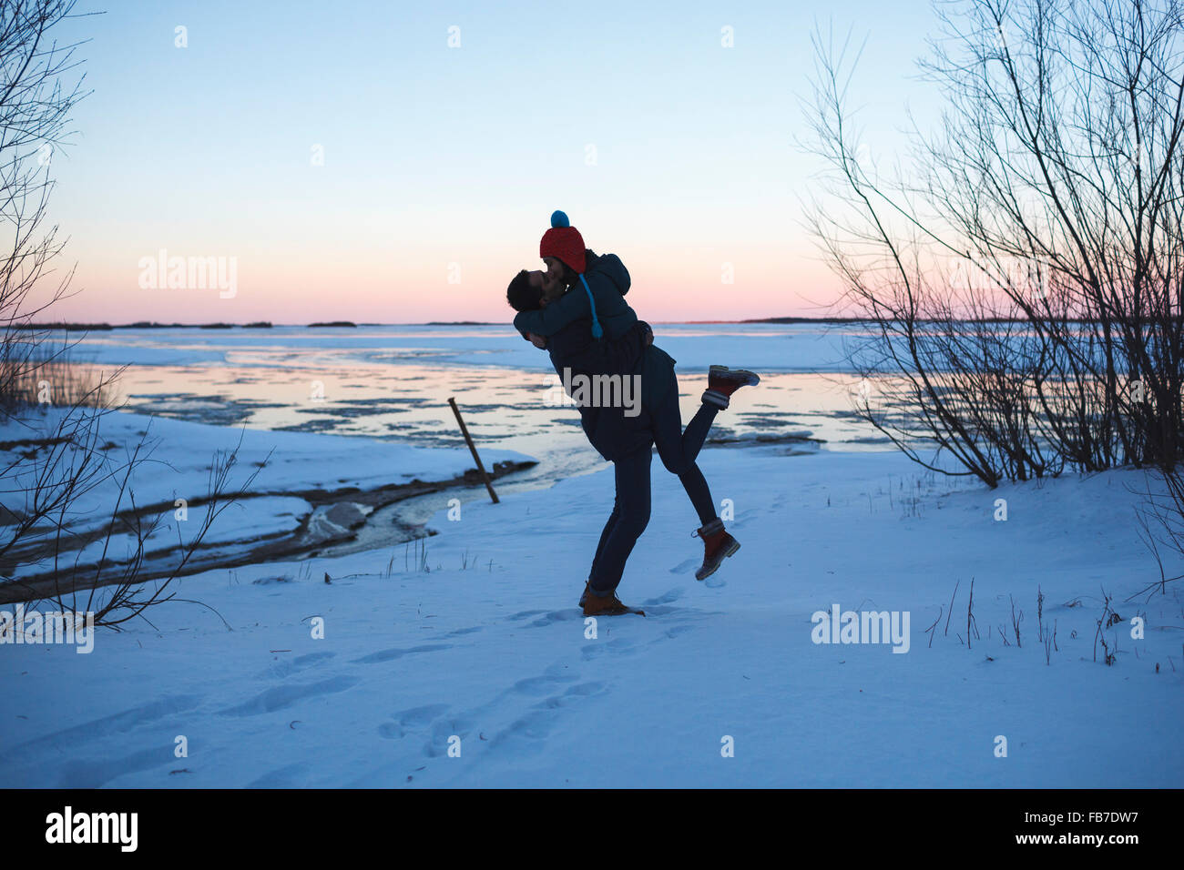 Full length of young man picking up woman and kissing on snow covered field Stock Photo