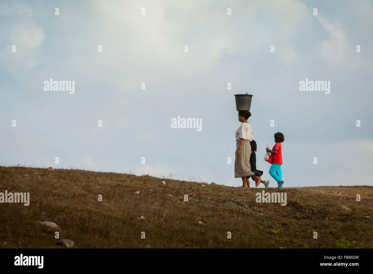 A woman carrying a plastic bucket on her head as she is walking with children on dry landscape during dry season in Sumba Island, Indonesia. Stock Photo