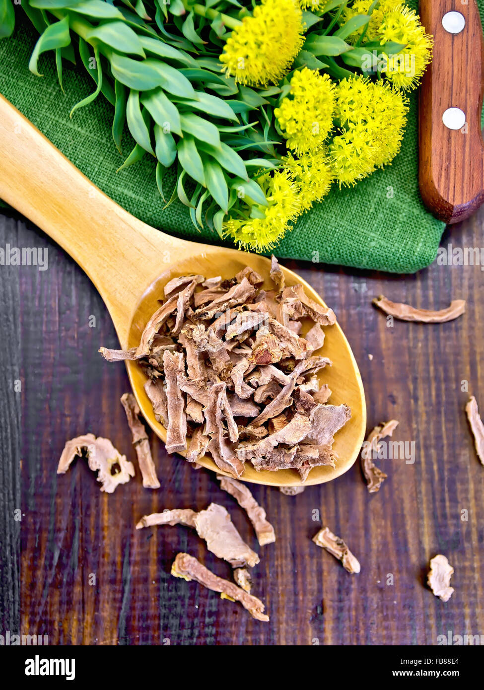 Spoon with dry roots of Rhodiola rosea, knife, fresh flowers Rhodiola rosea on a dark wooden board Stock Photo