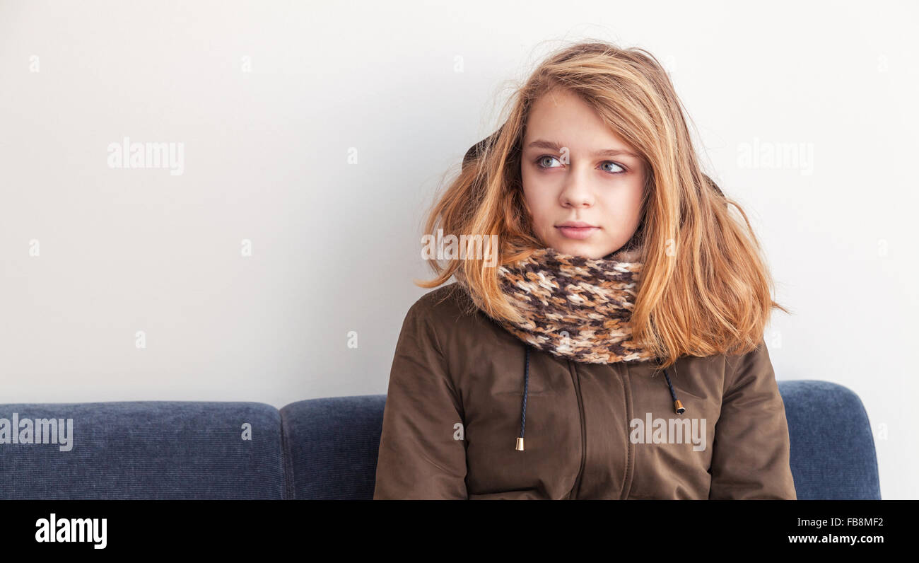 Beautiful blond Caucasian teenage girl in warm clothes sitting on blue sofa over white wall Stock Photo