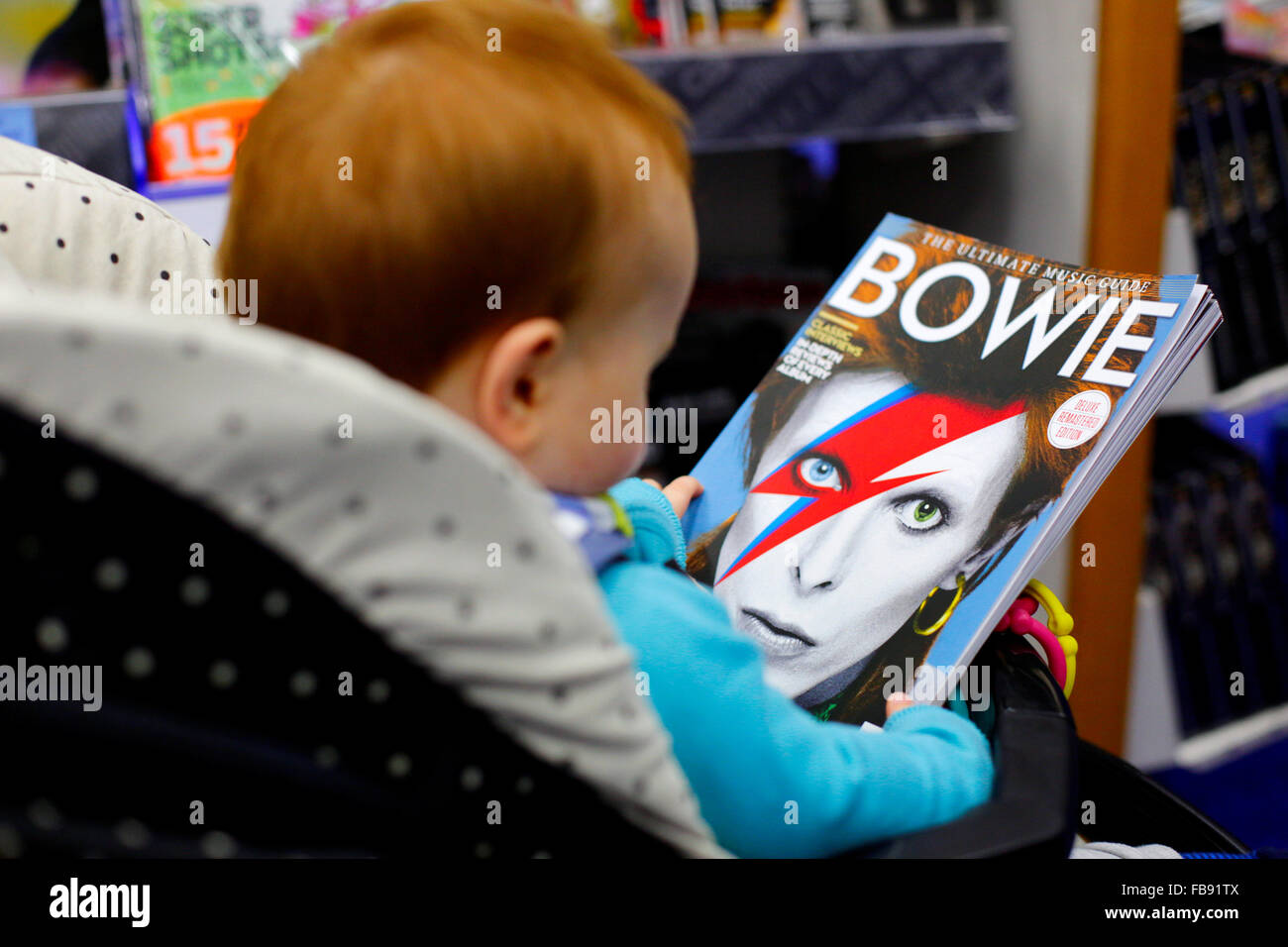 A young child in a pushchair, inside a shop, holding and looking at a magazine. The magazine is a bookazine tribute to David Bowie. The front cover of the publication has a full face portrait of Bowie with is distinctive lightening flash, made famous by his Aladdin Sane, alter ego character in the 1970’s. Stock Photo