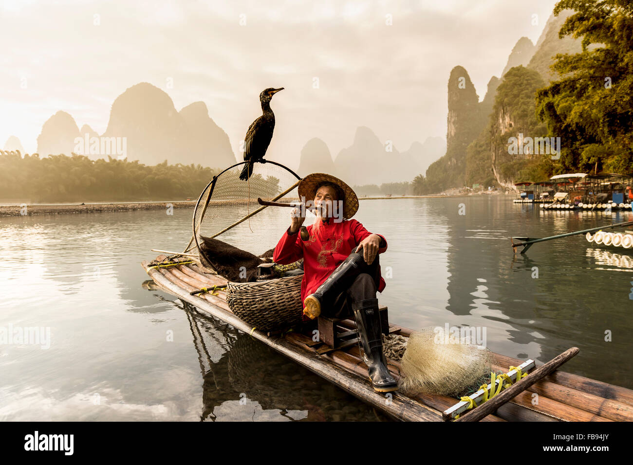 Li River - Xingping, China. Circa January 2016 - An old fisherman taking a smoke break with his cormorant on a bamboo raft. Stock Photo