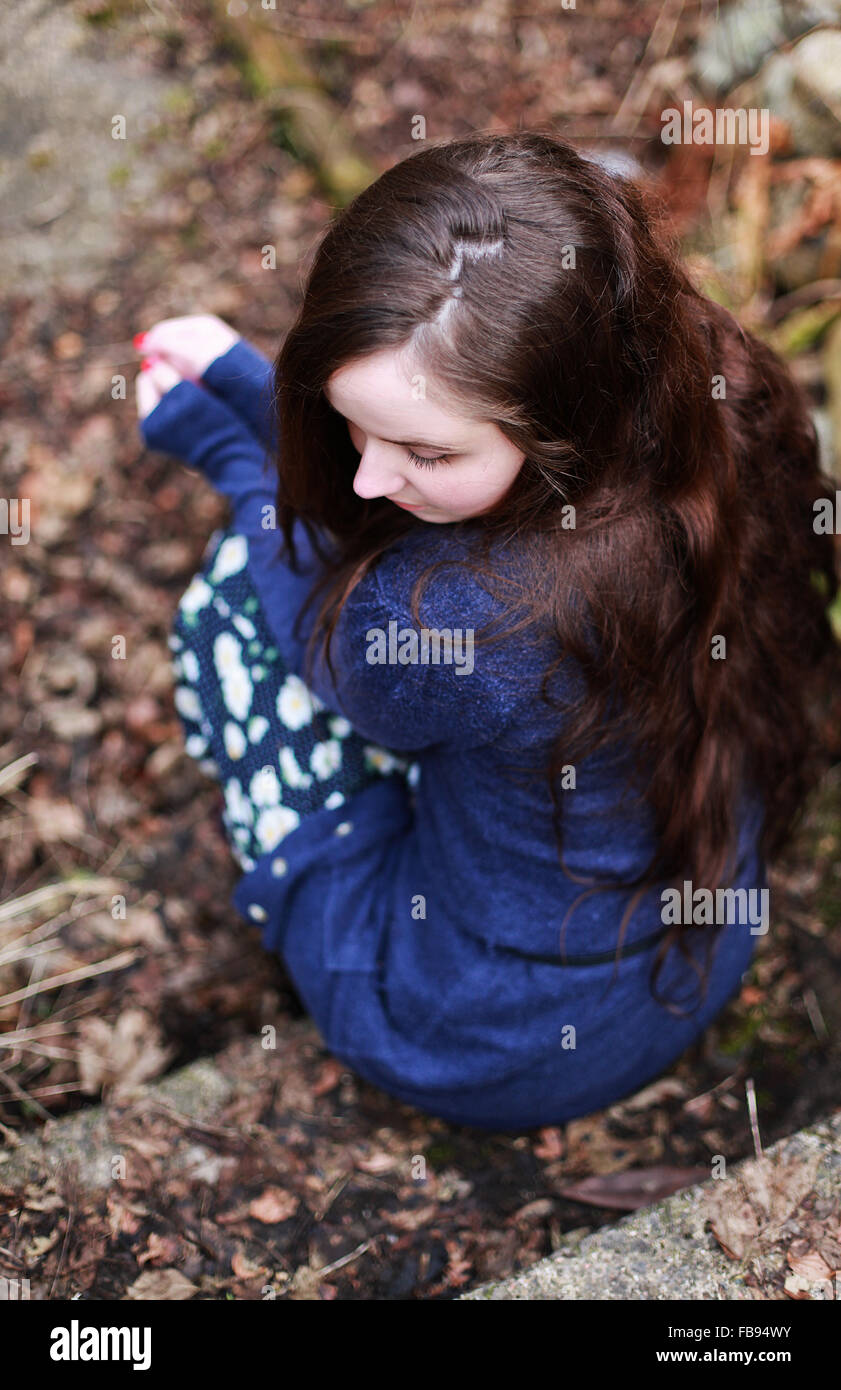 Back of a young woman with long brown wavy hair sitting on a step waiting Stock Photo