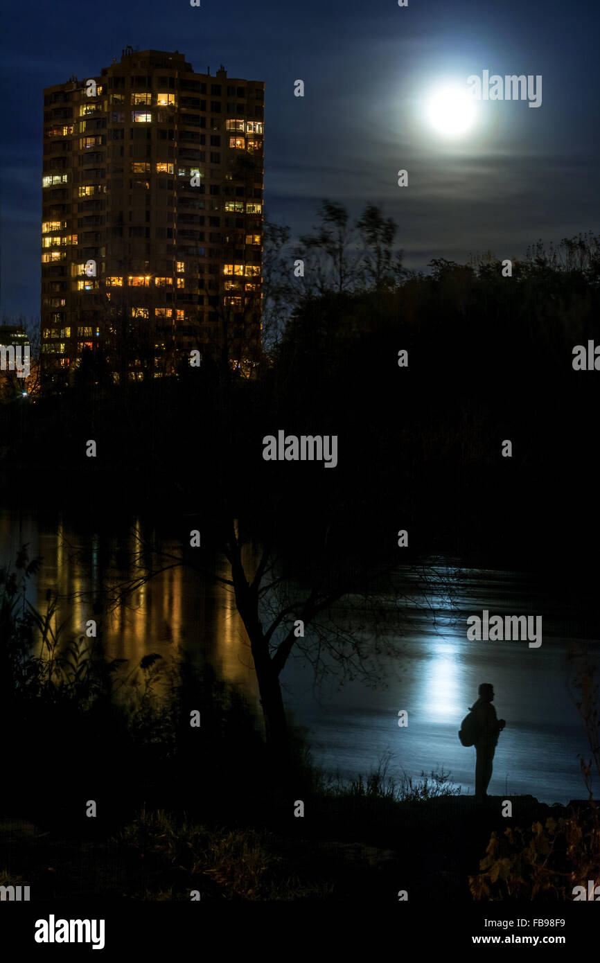 a unidentifiable man is standing on the bank of a river in the moolight reflexion, there is a condo building reflecting in the w Stock Photo