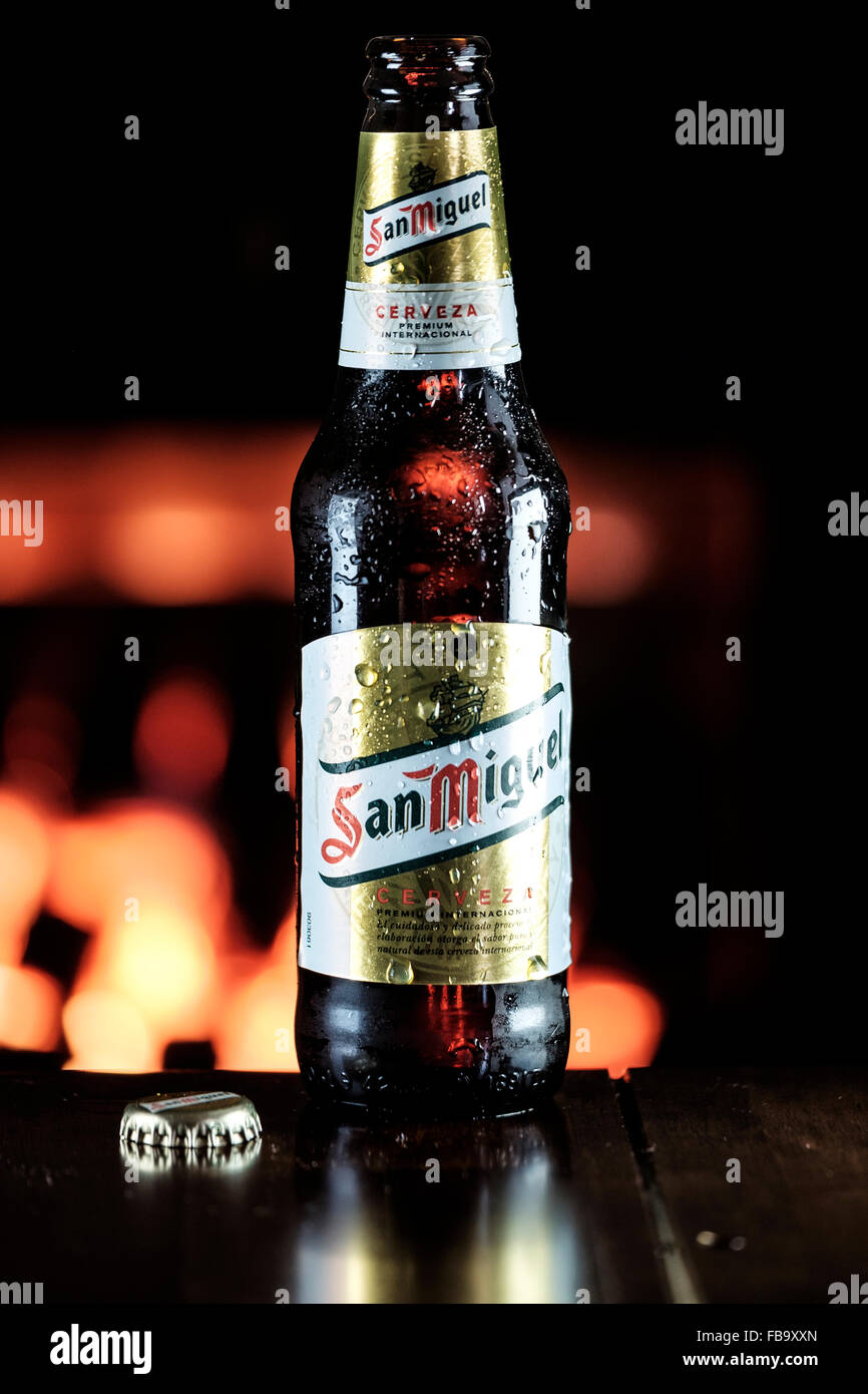 A cold bottle of San Miguel beer on a wooden table in front of a warm, glowing fire. Stock Photo