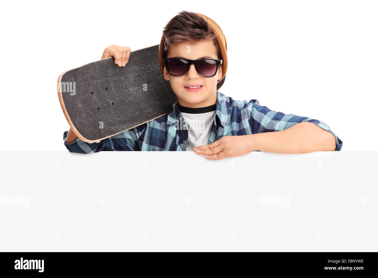 Cool little boy with sunglasses holding a skateboard and posing behind a signboard isolated on white background Stock Photo
