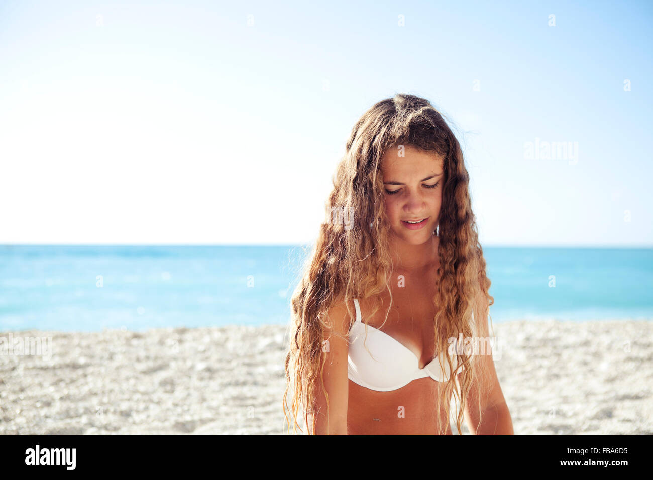 Young girl teenager looking down on a white beach with light blue ocean behind. Stock Photo