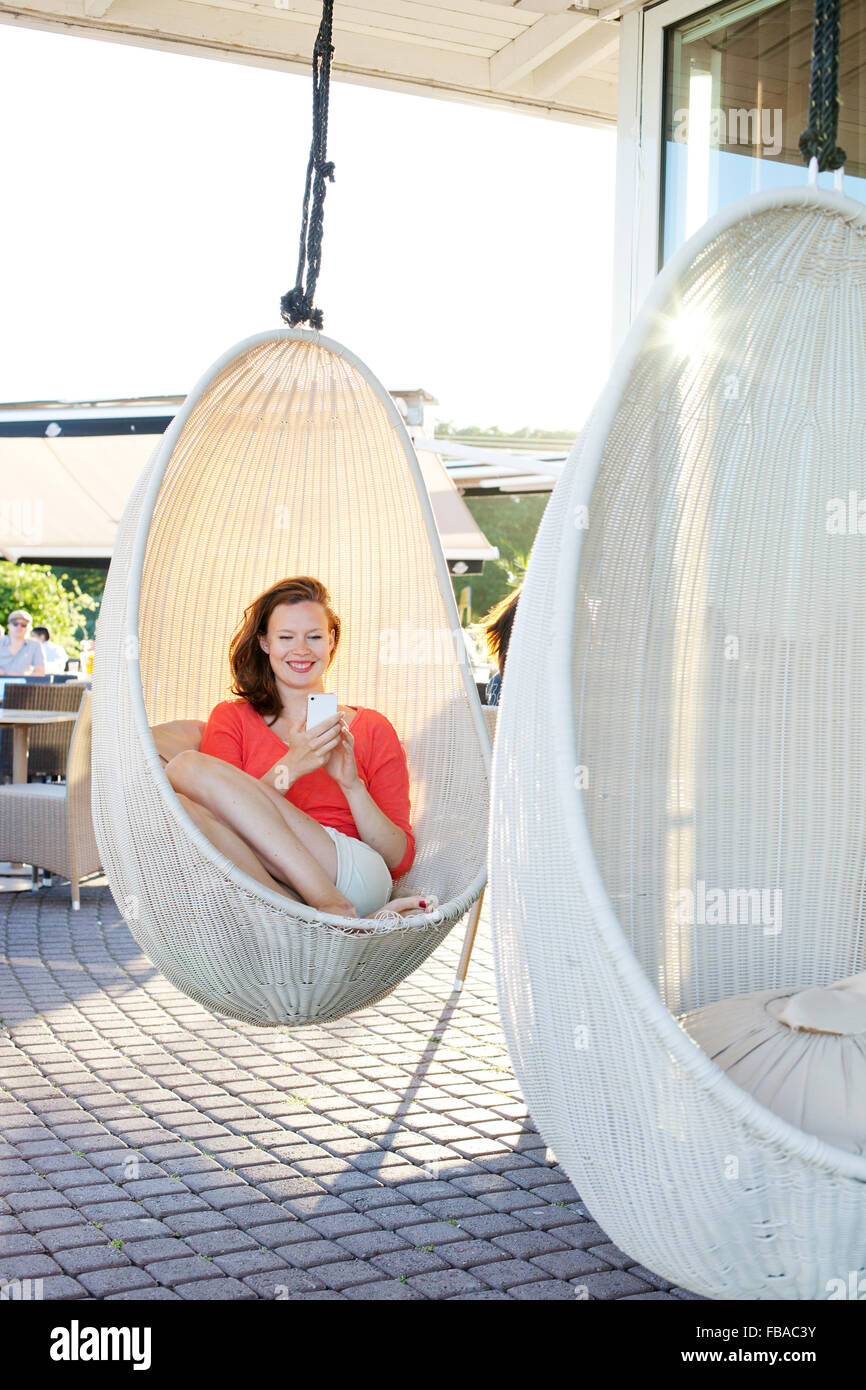 Finland, Uusimaa, Helsinki, Kaivopuisto, Smiling young woman using smart phone in swing chair Stock Photo