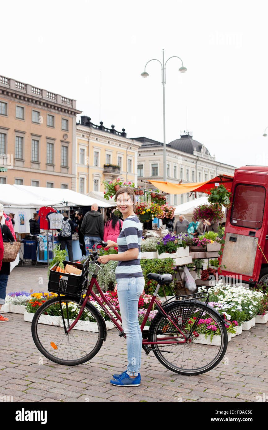 Finland, Uusimaa, Helsinki, Kauppatori, Portrait of smiling young woman with bicycle at street market Stock Photo