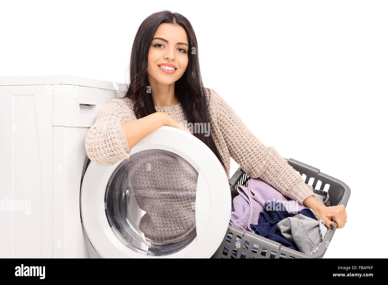 Young cheerful housewife holding a laundry basket and posing next to a washing machine isolated on white background Stock Photo