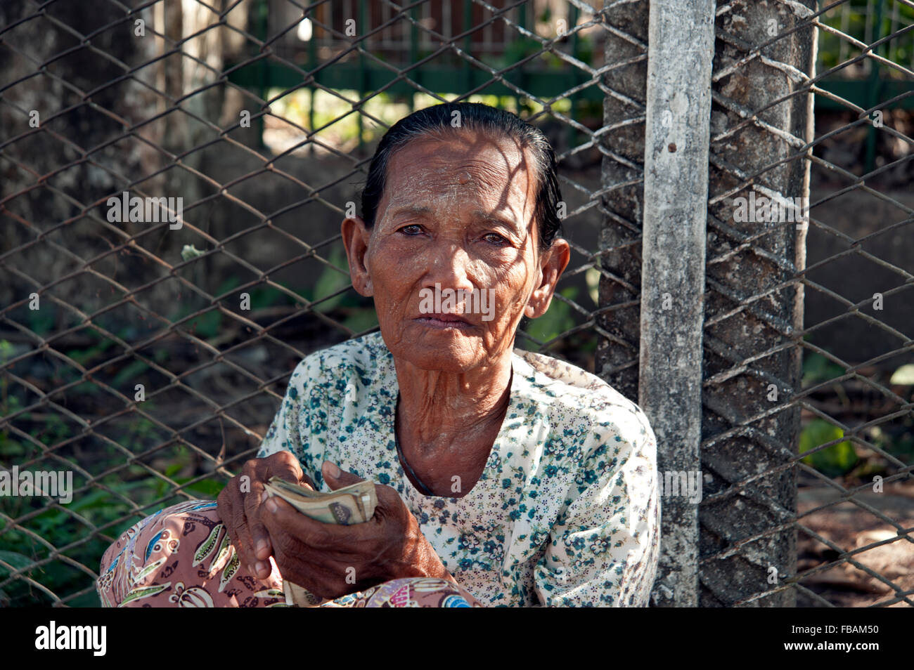 An old Burmese lady begging sitting on the pavement against a wire fence on a Rangoon street Myanmar Burma Stock Photo