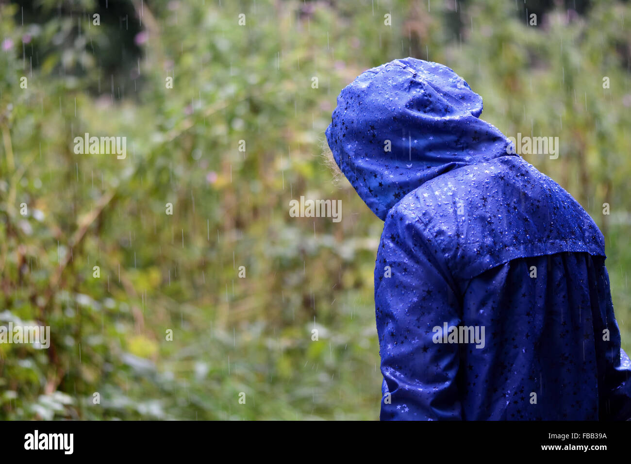Child in a blue waterproof coat in the rain. A girl stands with her head bowed in heavy rain, in a bright blue raincoat Stock Photo