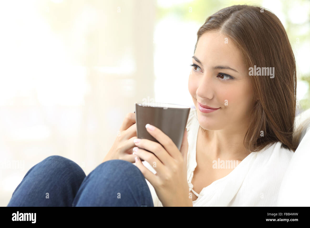 Pensive candid girl looking sideways and holding a coffee cup sitting on a couch at home Stock Photo