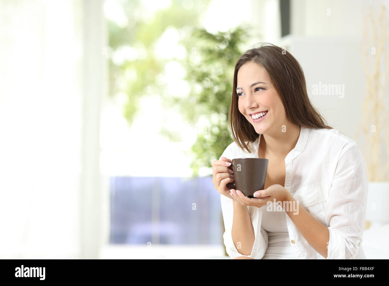 Pensive woman drinking coffee or tea and thinking looking sideways at home Stock Photo