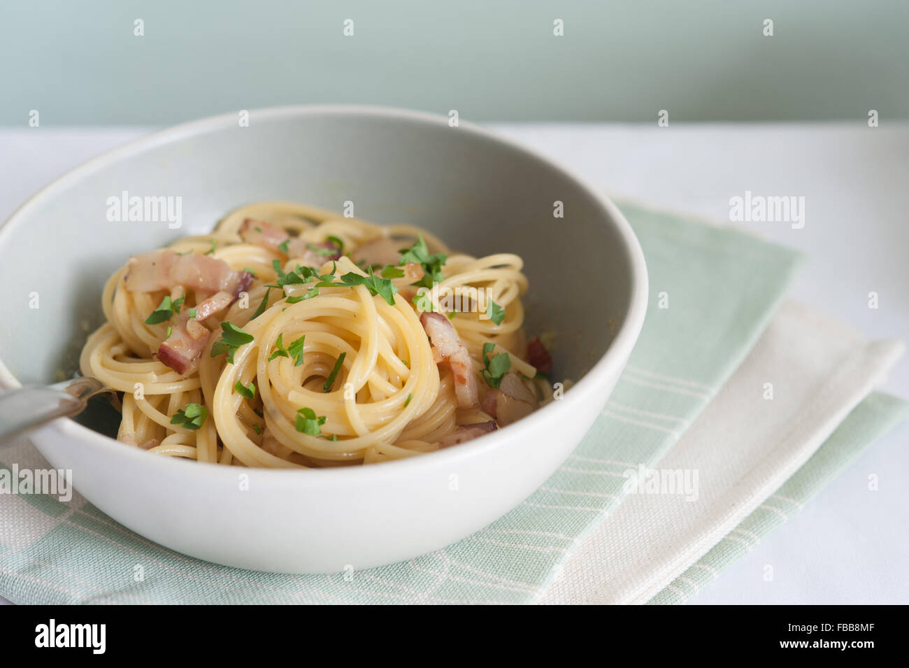 Spaghetti pasta with bacon in light grey and white bowl garnished with parsley on pastel linens and light grey background Stock Photo