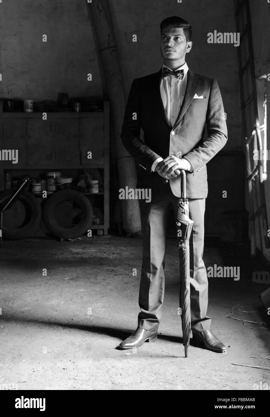 Black and White classic portrait of a young man dressed in black suit, bow tie, posing with umbrella in an old garage room. Stock Photo