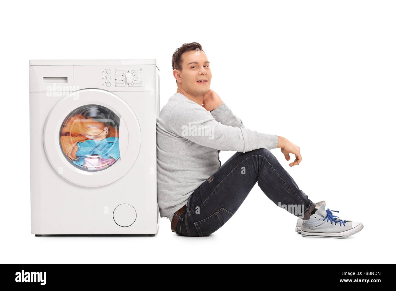 Young man sitting next to a washing machine and waiting for the laundry isolated on white background Stock Photo