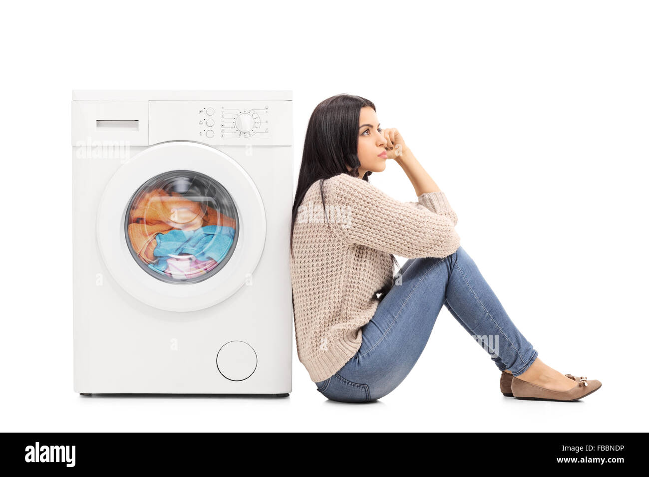 Young pensive housewife sitting on the floor waiting for the washing machine to finish isolated on white background Stock Photo