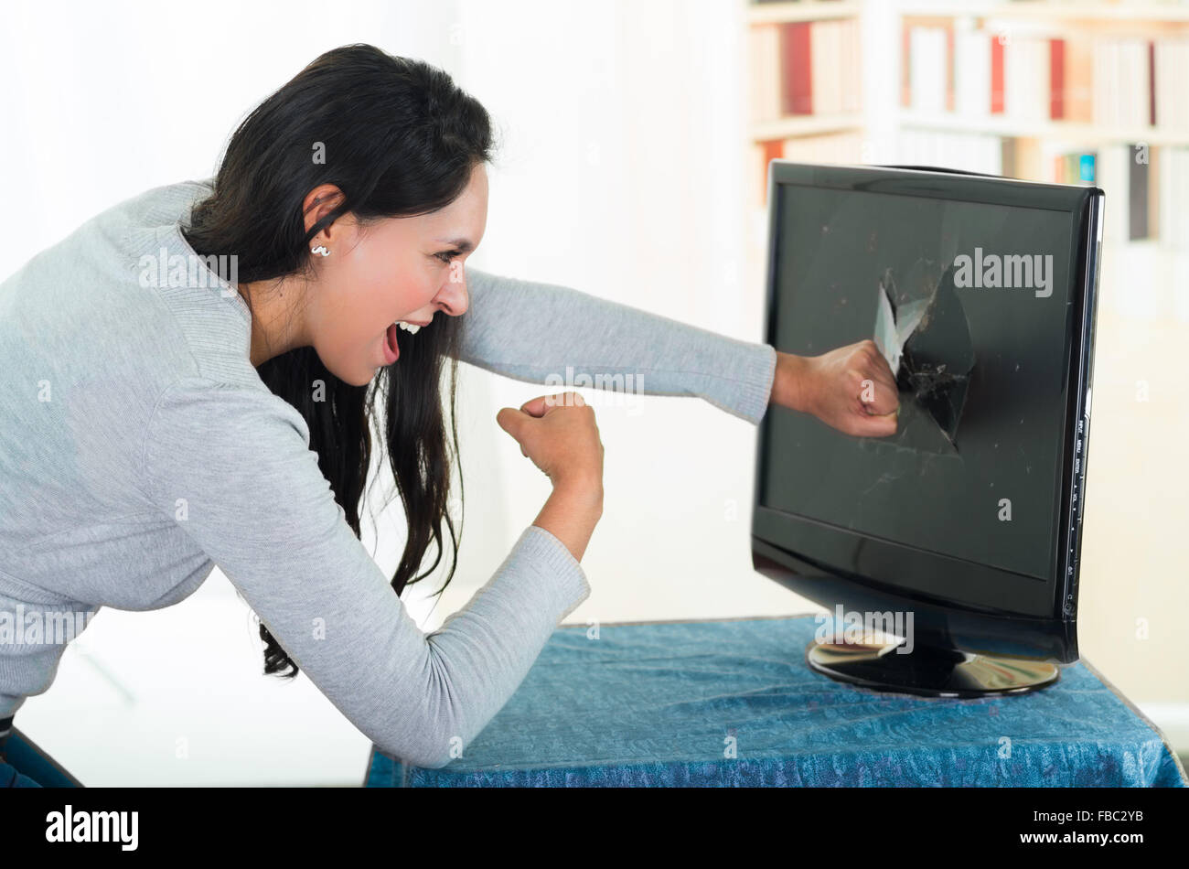 Angry female brunette punching computer screen with large hole on it Stock Photo