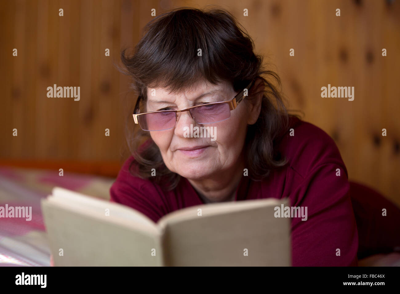 Cute senior woman in glasses reading a book, lying comfortable on bed