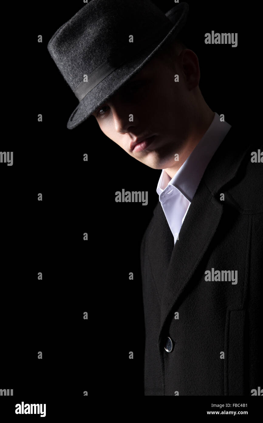 Handsome brutal looking man in coat and hat in the darkness, studio shot against black background, low key lighting Stock Photo