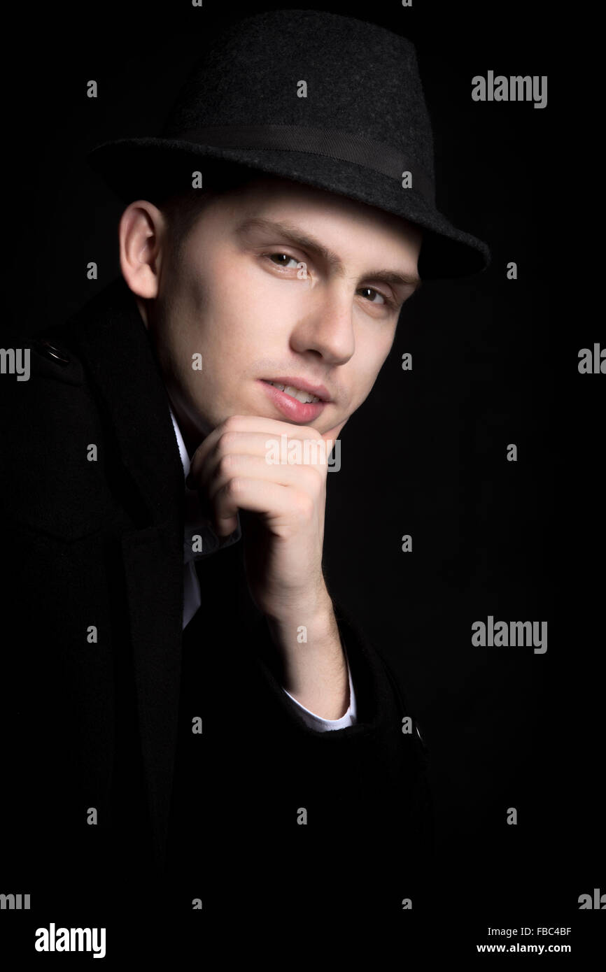 Portrait of smiling good-looking young man in hat and coat with confident look, studio shot against black background Stock Photo