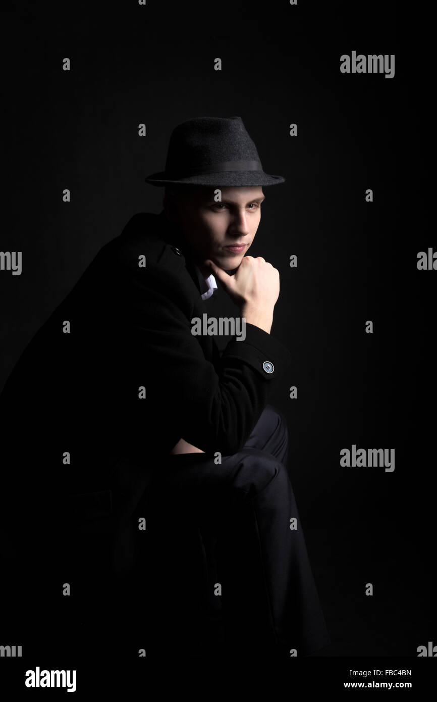 Low key shot of mysterious young man wearing hat and coat sitting in the darkness with thoughtful look Stock Photo