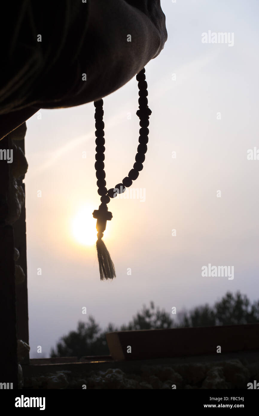 Silhouette of prayer hands holding Christian rosary with cross against the sun in the sky Stock Photo