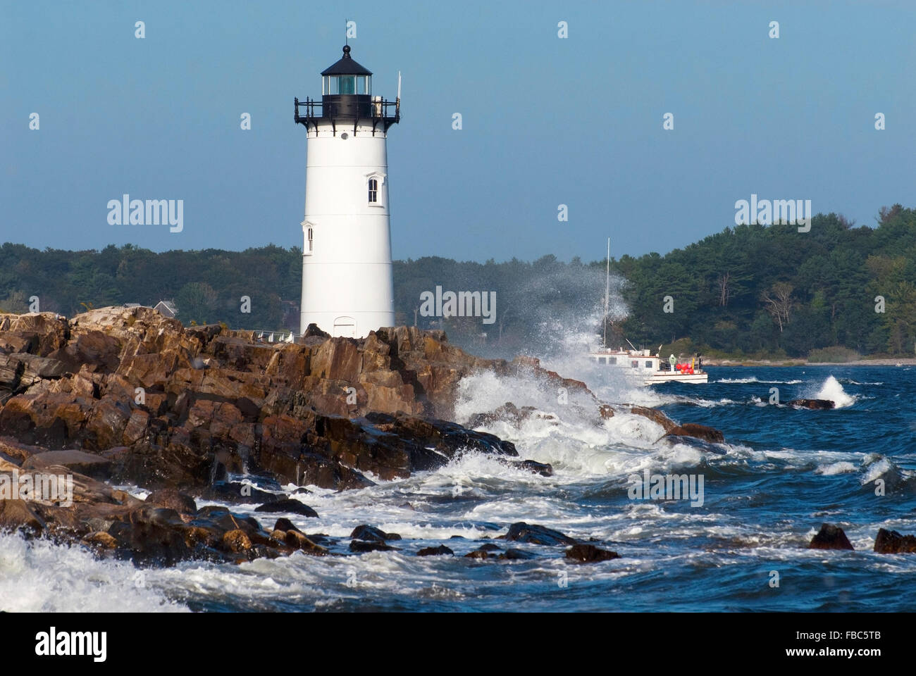 Portsmouth Harbor lighthouse guides fishing boat through rough surf along rocky shore of New Hampshire. Stock Photo