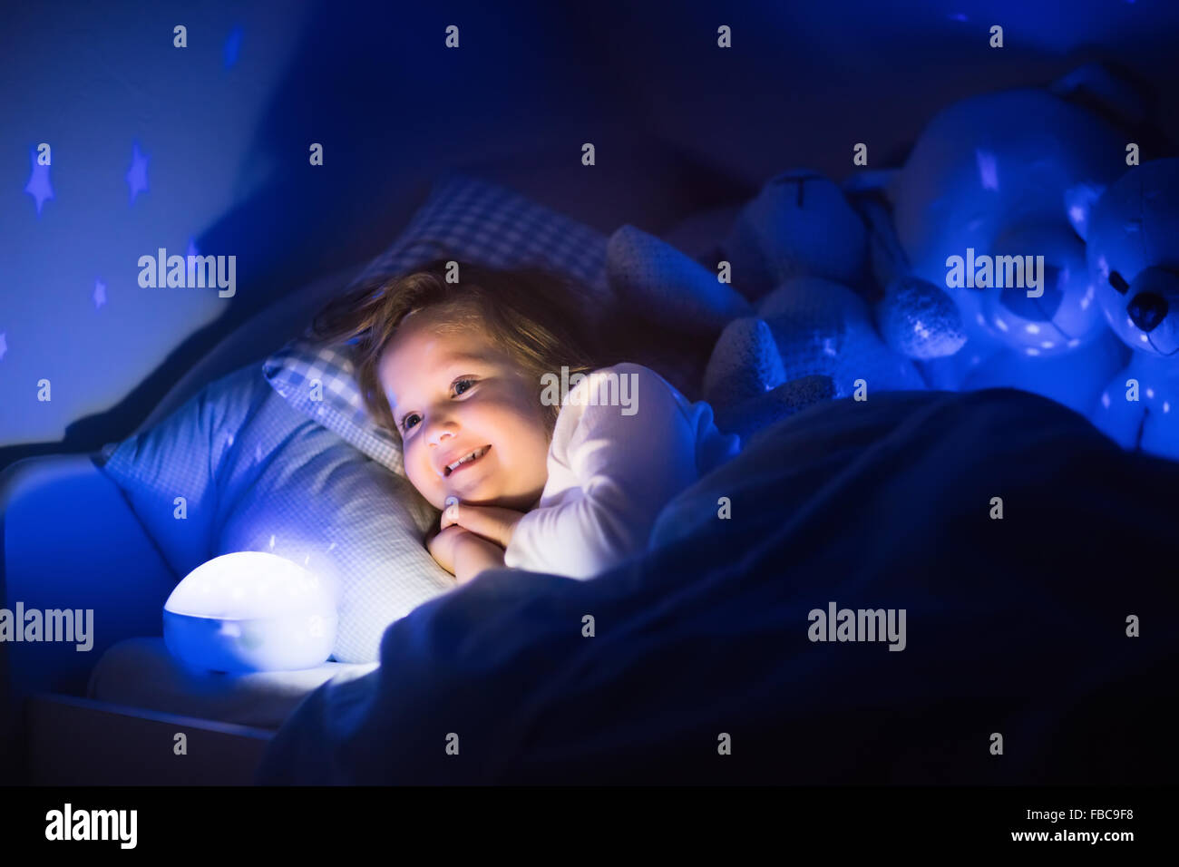 Little girl reading a book in bed. Dark bedroom with night light projecting stars on room ceiling. Kids nursery and bedding. Stock Photo