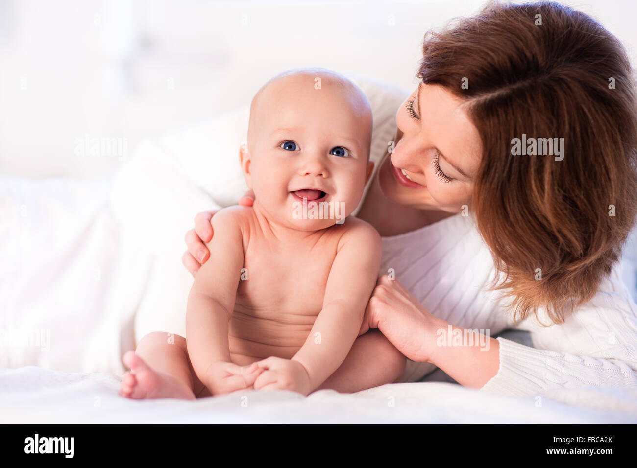Mother and child on a white bed. Mom and baby boy in diaper playing in sunny bedroom. Parent and little kid relaxing at home Stock Photo