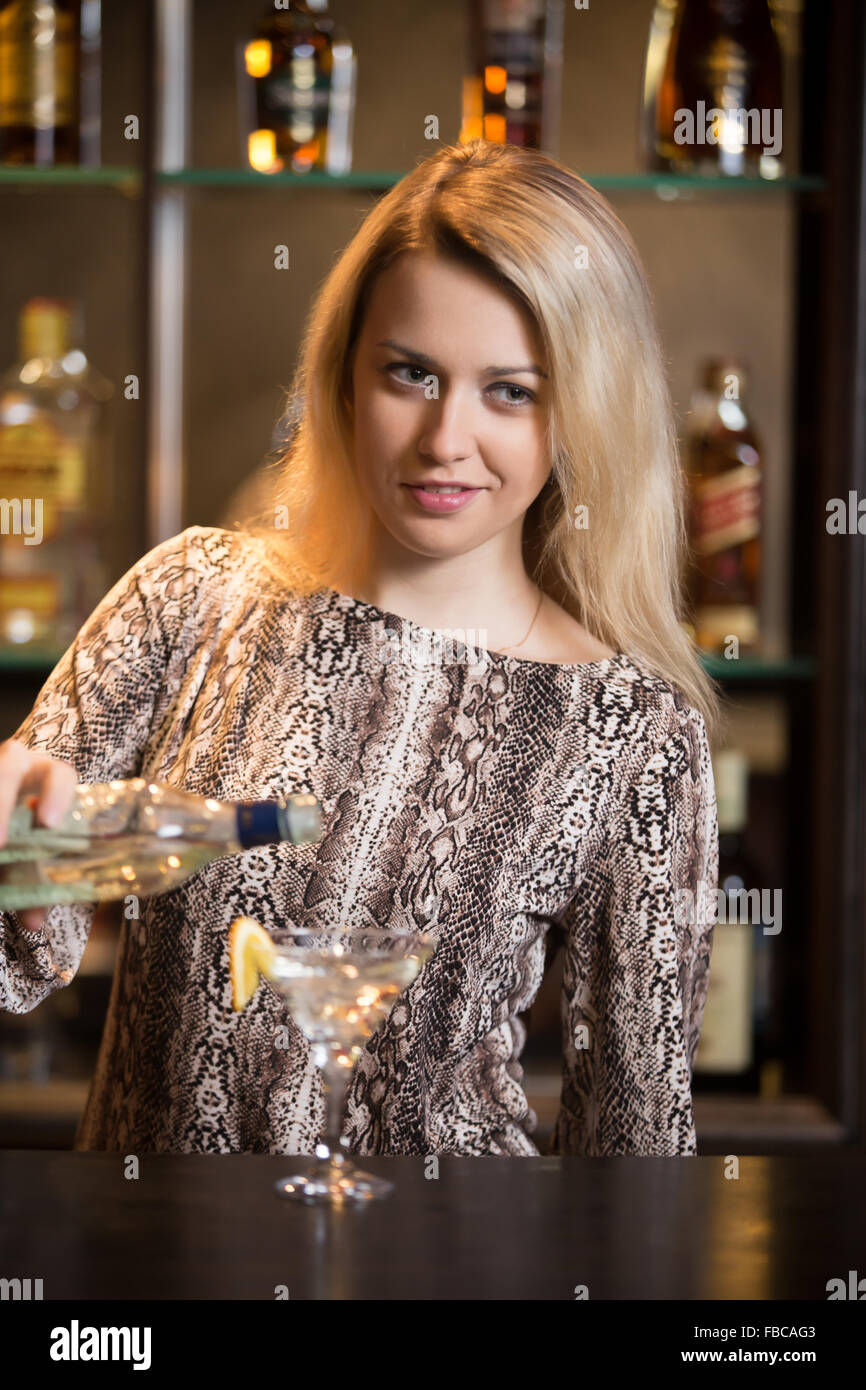 Working blond female bartender, making cocktail at bar counter, holding bottle in hand, pouring drink in martini glass Stock Photo