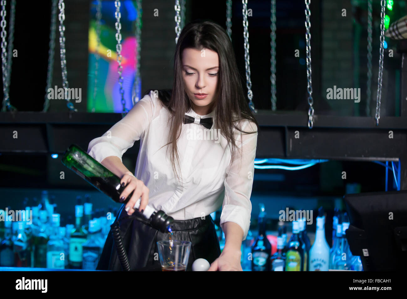 Beautiful brunette bartender girl wearing white shirt and black bow tie, serving alcohol drink at bar counter, holding bottle in Stock Photo