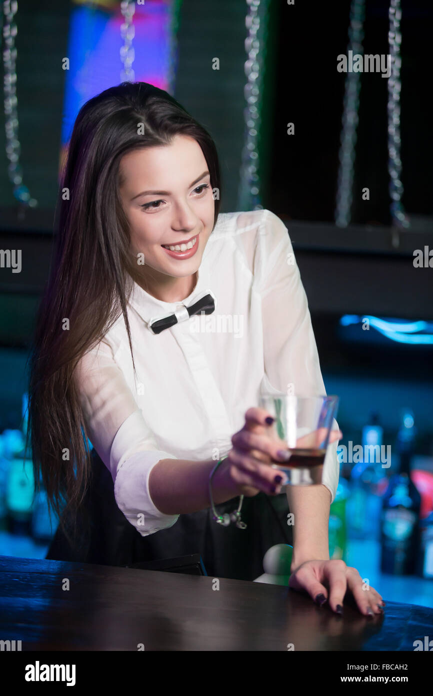 Beautiful cheerful brunette bartender girl in white shirt and black bow tie, serving alcohol drink at nightclub bar, holding gla Stock Photo