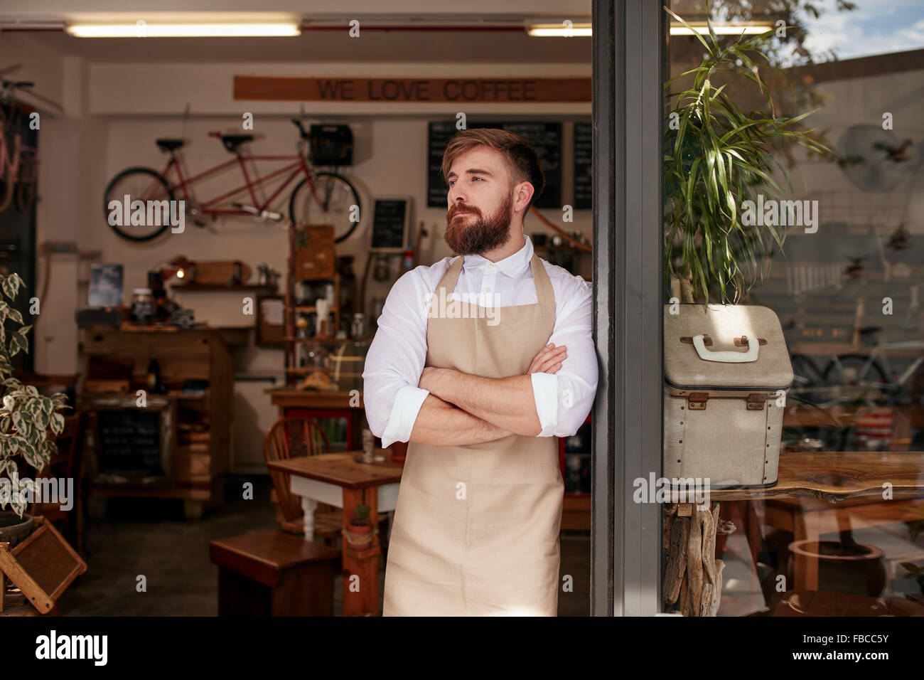 Portrait of a cafe owner standing in the doorway of his coffee shop. Young waiter standing with his arms crossed at the door of Stock Photo